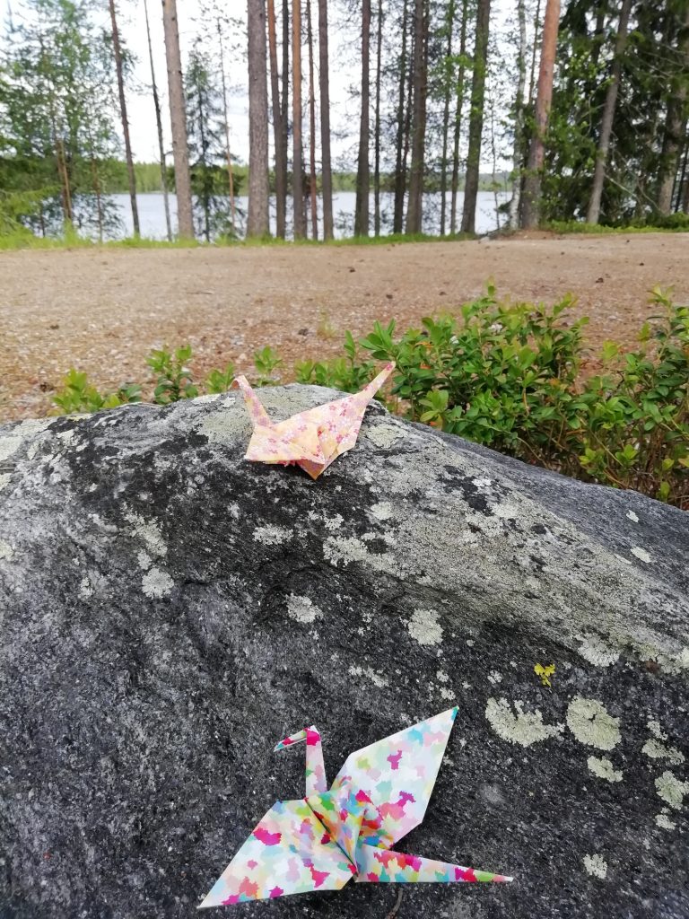 Kaksi origamilintua kivellä. Taustalla näkyy järvi.